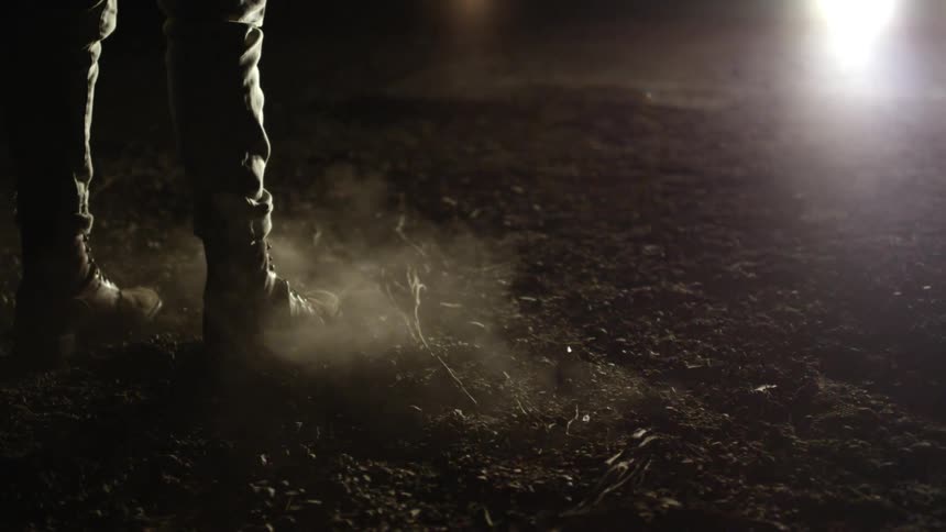 Person standing and kicking dirt in the dark while headlights shine toward them