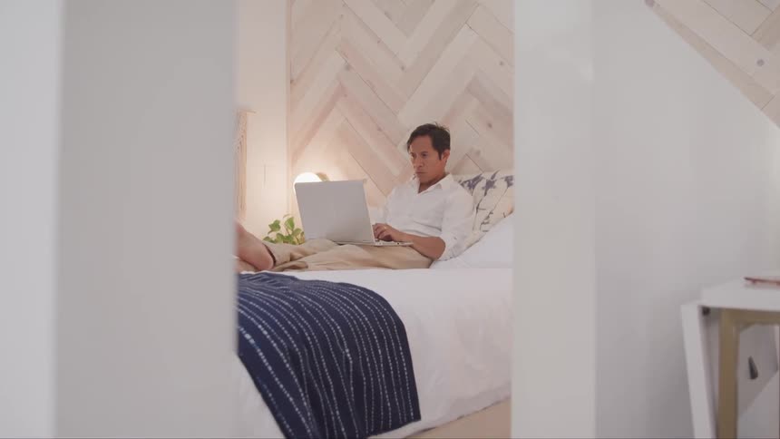 A man sitting up in bed with legs stretched out and crossed working on a laptop