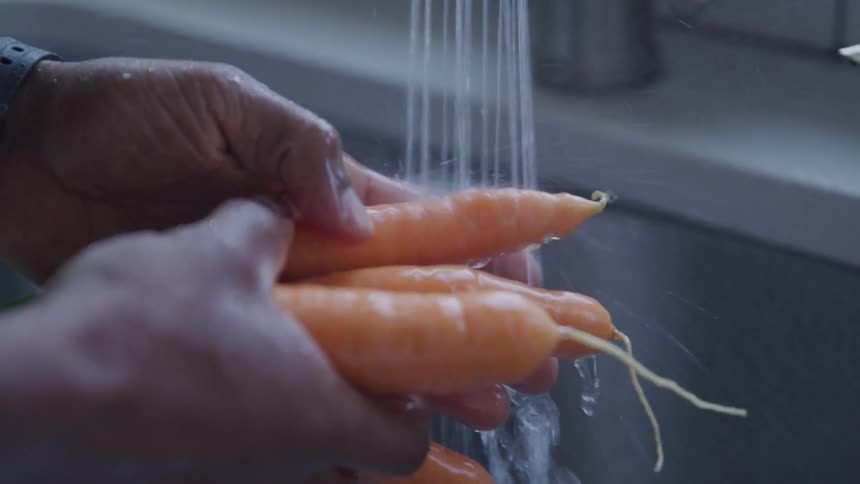 Hands hold and wash carrots under the water spray from a kitchen faucet