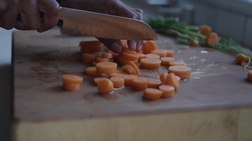 A person holds a sharp stainless steel knife as they cut sliced carrots in half on a cutting board