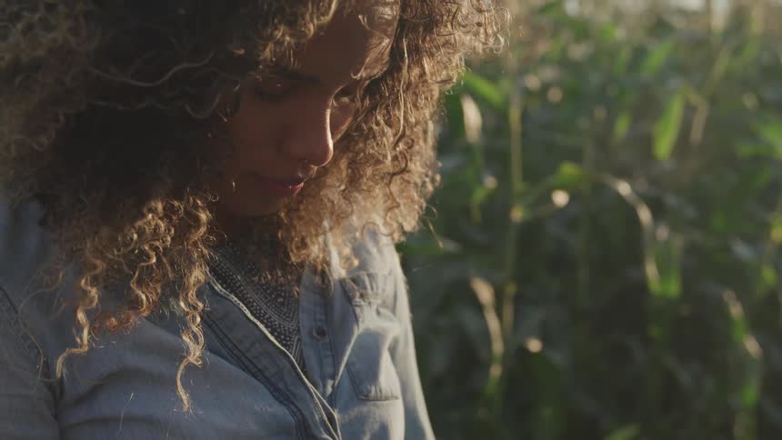 Curly haired brunette woman with head down stands on the farm path in the corn field