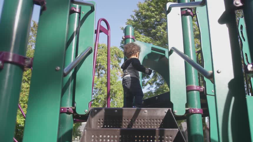 A toddler climbs up the last step to a slide in the playground on a sunny day