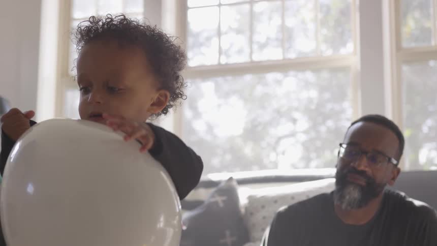 Black man sitting on the ground looks at the toddler boy who plays with a white balloon in the living room
