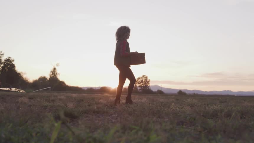 Brunette woman holding a box walks on dry grass at sunset