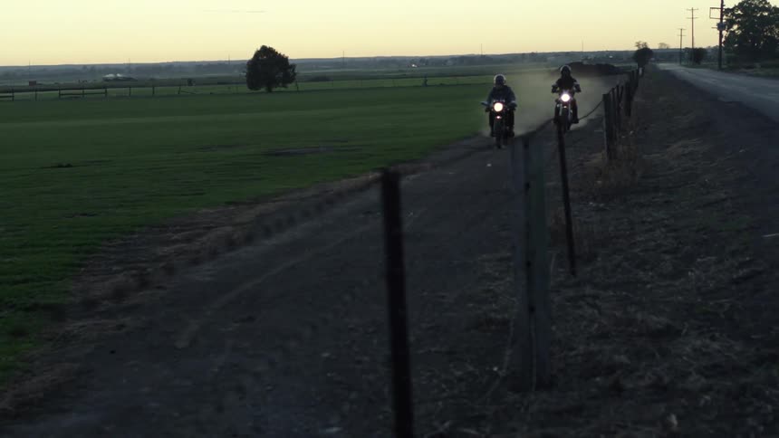 Two motorcyclists ride on path beside fence