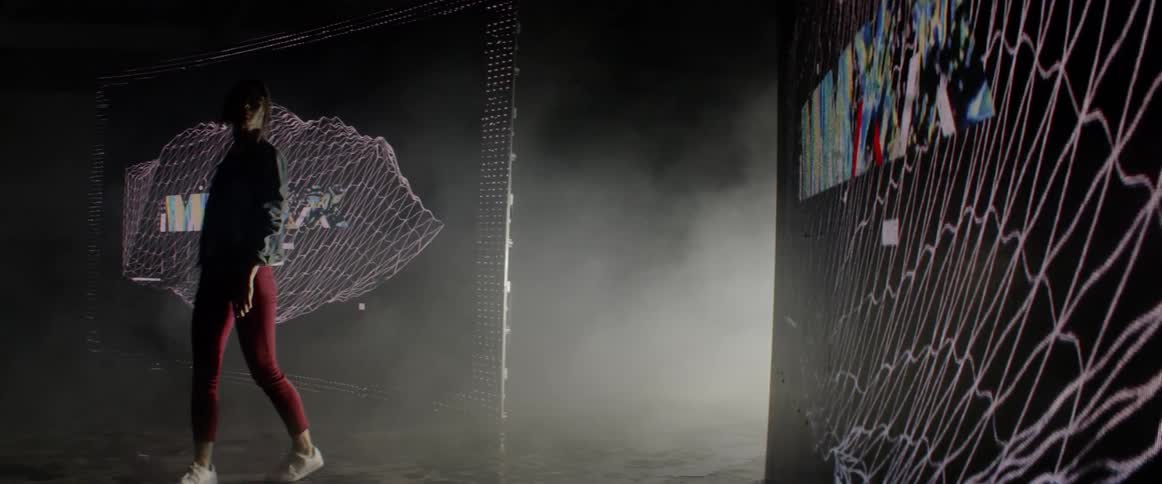 A woman walks between two large screens then turns around and looks at them
