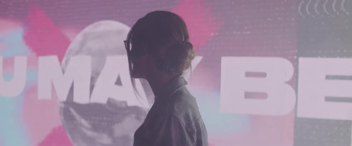 A woman stands in front of a large video screen with words running across it