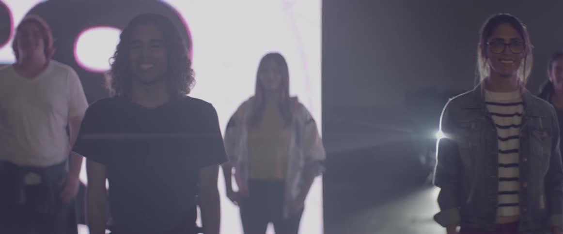 Young women and men stand on a stage smiling in front of a digital display showing large numbers