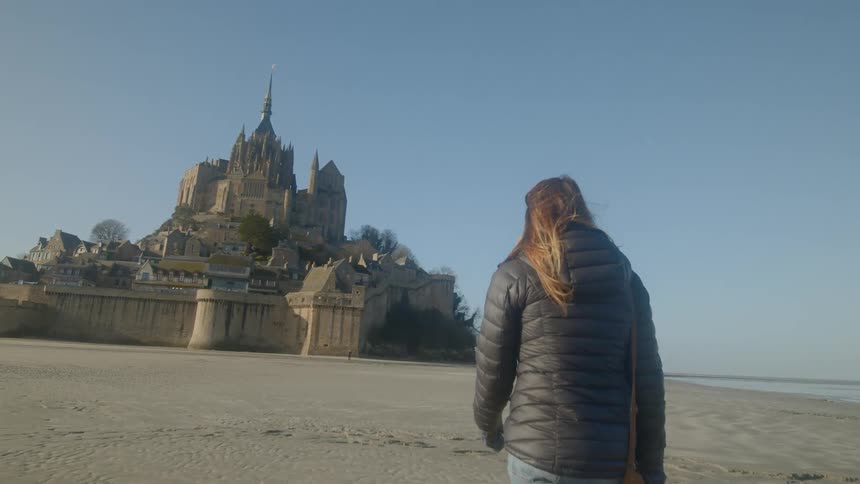 A woman in a waterproof coat walks towards a large period building on a coast