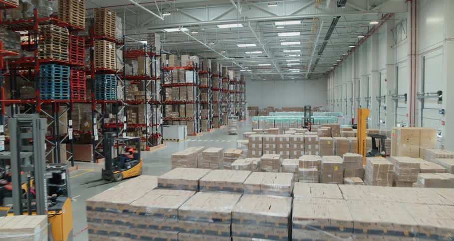 Forklifts move pallets of boxed goods that sit next to the bay doors of a warehouse