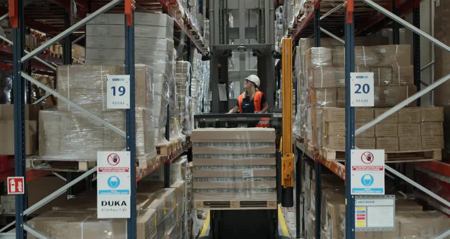 A woman drives a forklift of pallets to a high shelf