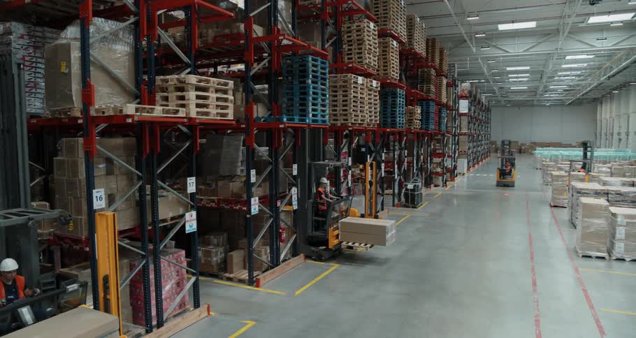Within a massive warehouse multiple forklifts are being used to move pallets of supplies to tower shelves that are stacked high