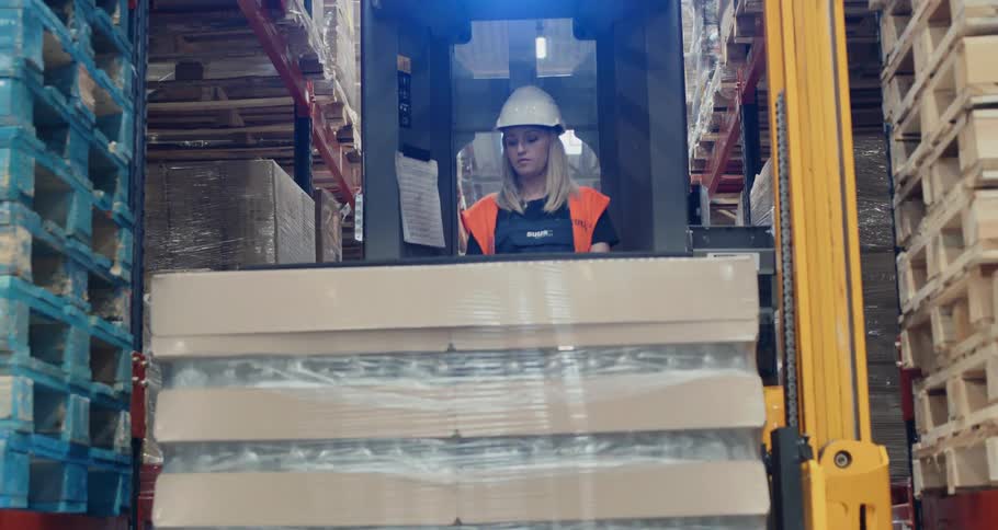 A woman is lowering a pallet of goods in the aisle of a warehouse