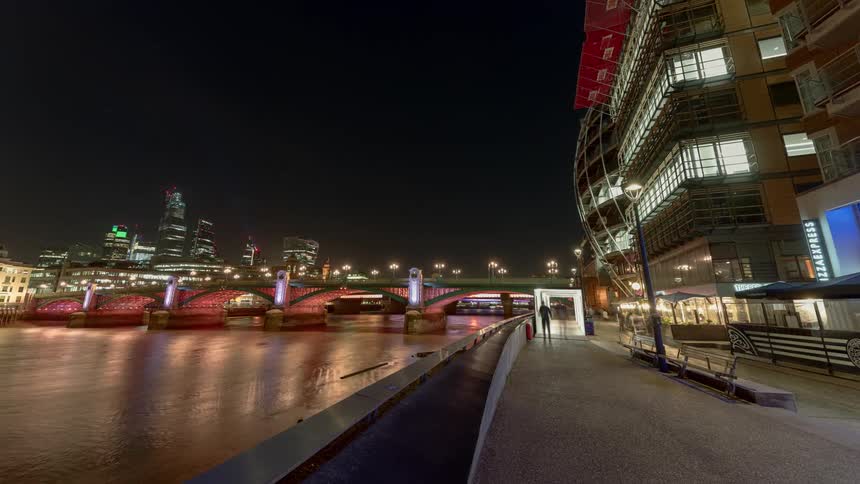 People sit and walk along the citys waterfront as cars travel across the bridge at night