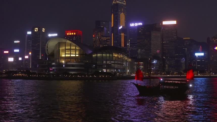 A red sail junk boat travels past the hong kong skyline at night