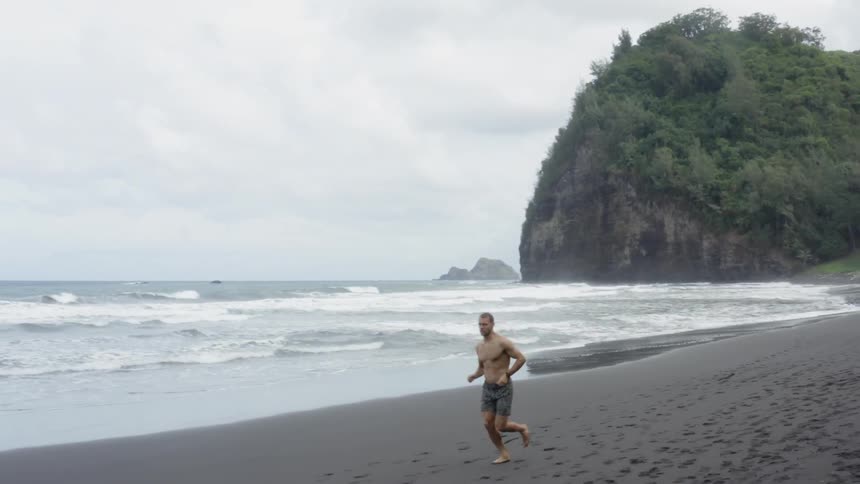 Shirtless man runs on the beach next to the ocean on a cloudy day