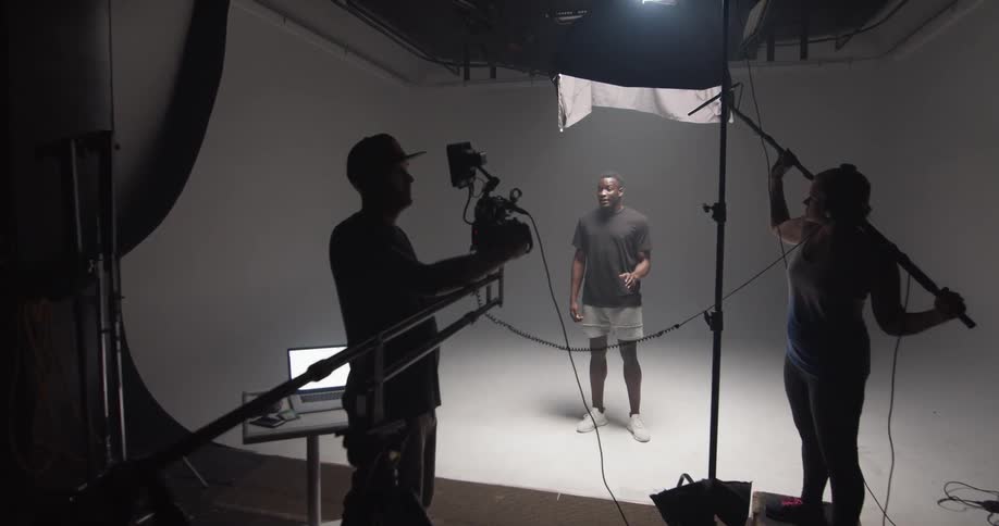 A man stands alone on a soundstage and talks while be filmed and recorded