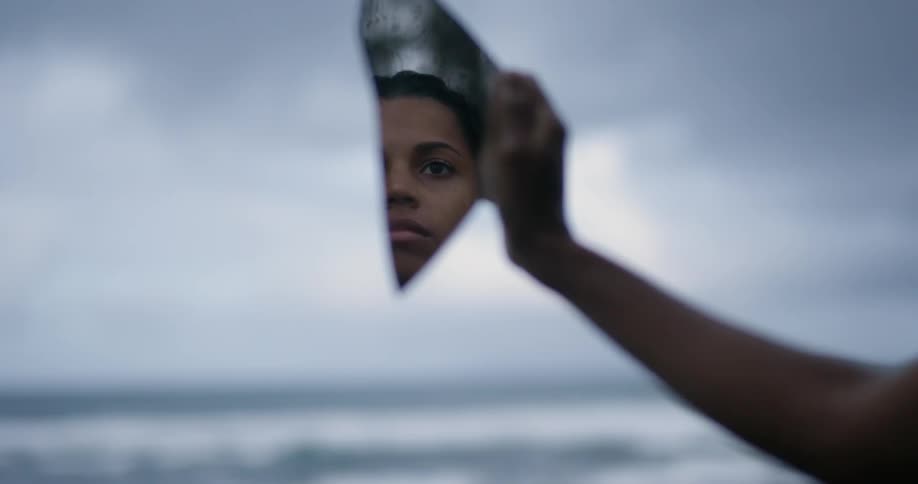 Woman looks at her reflection in a piece of broken mirror while near the ocean