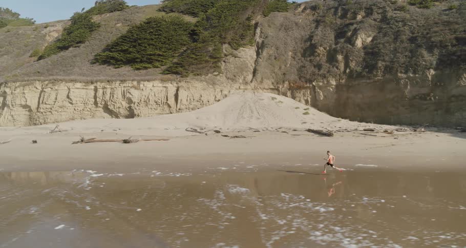 A man jogs along the water edge with hills on the other side of him