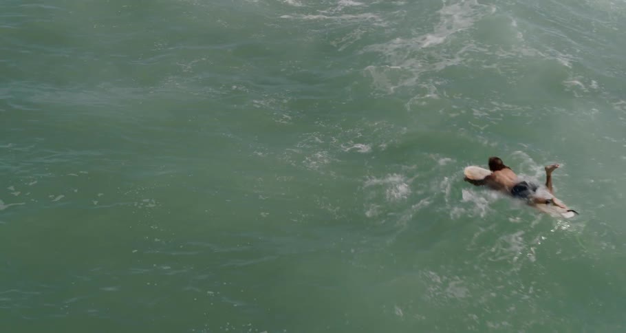 Male lays on a surfboard and paddles with his arms out into the ocean
