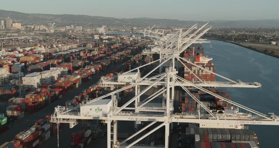 A river is lined with multiple large cranes and maritime shipping yards that are packed with cargo containers