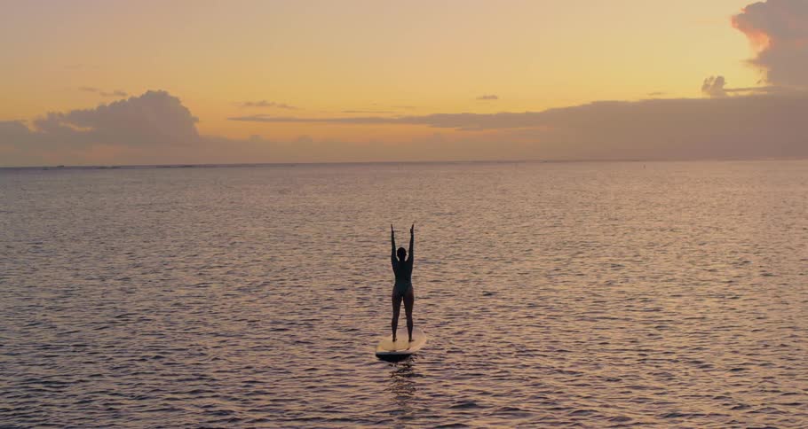 A woman standing on a large surfboard that is floating on the ocean stretches her arms above her head