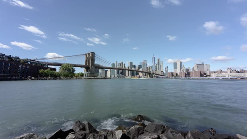 Water splashes against shore as boats pass under suspension bridge near city downtown