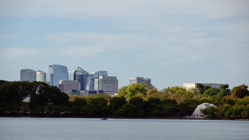 People walk along the water as a boat travels in the river with a city skyline behind