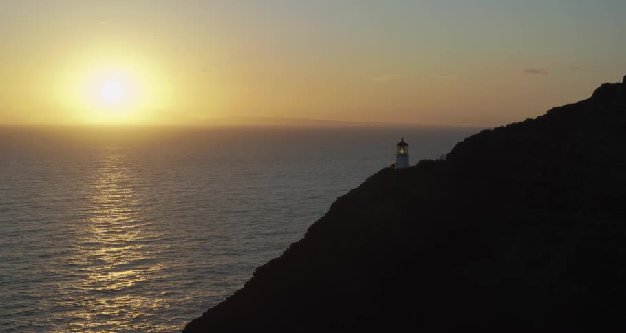 A lighthouse sits on a hill overlooking the water and the setting sun