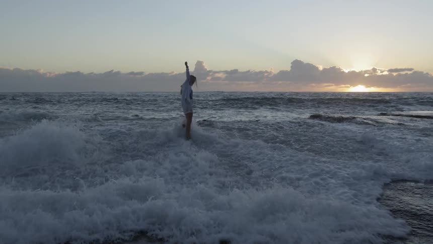 A young woman walks up to a rocky shoreline as a wave crashes into her