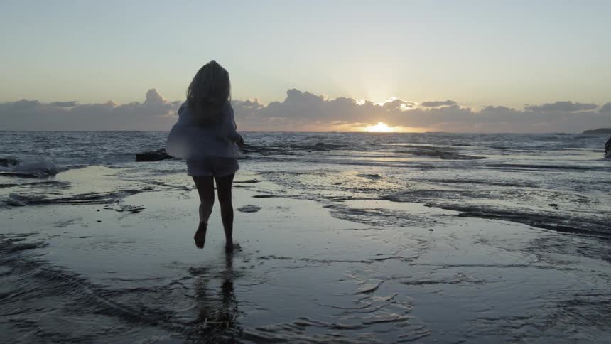 A woman in a light colored shirt runs along the beach at sunset