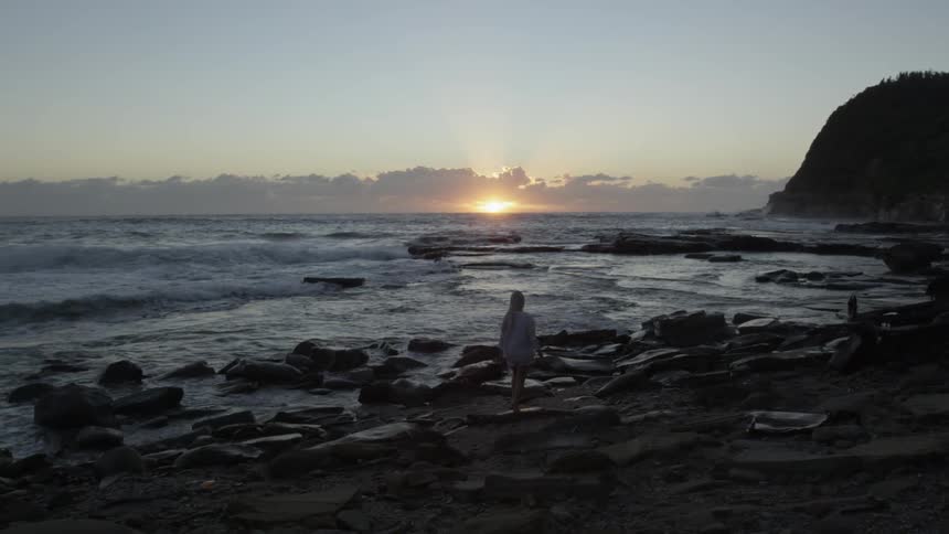 A blonde woman walks along a rocky shoreline as the sun sets