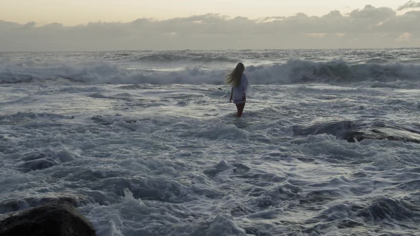 A blonde woman wading in the rough surf on a cloudy day