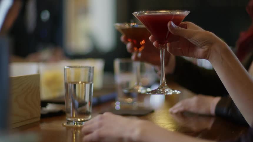 A man and a woman sitting at a bar pick up martini glasses and toast