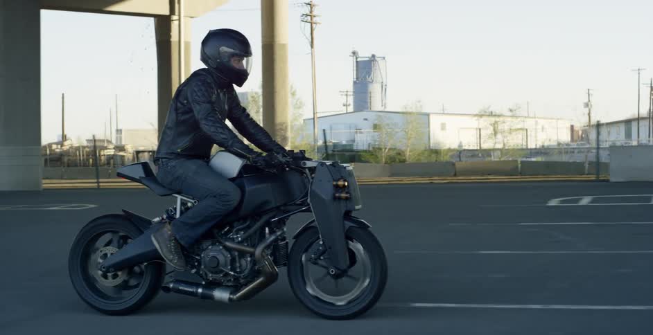 A man rides a motorcycle through a parking lot under an overpass