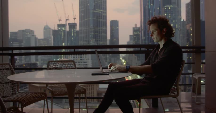 A man sits at a table while typing on his laptop and looking out his window at the city skyline outside