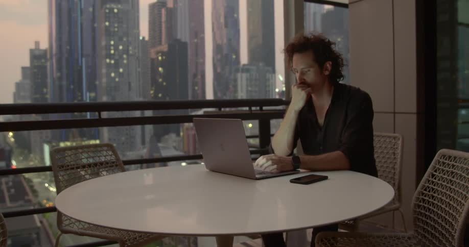 A man with curly hair sits at a table while typing on his laptop and numerous skyscrapers are outside his window
