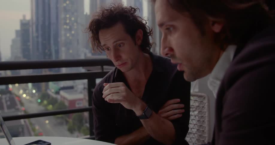 Two men talk while working on a laptop with skyscrapers outside the window