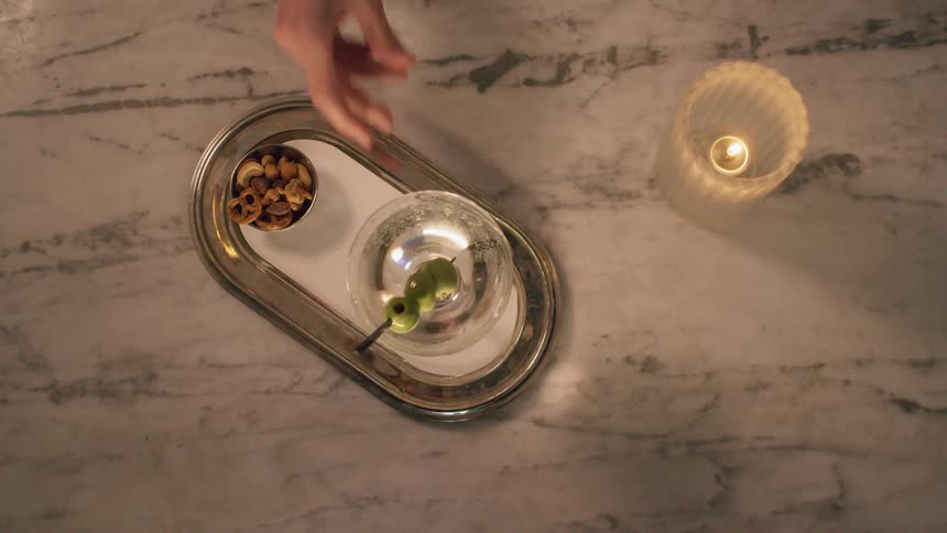 A martini with three olive garnish is placed on a tray next to a small bowl of nuts and pretzels
