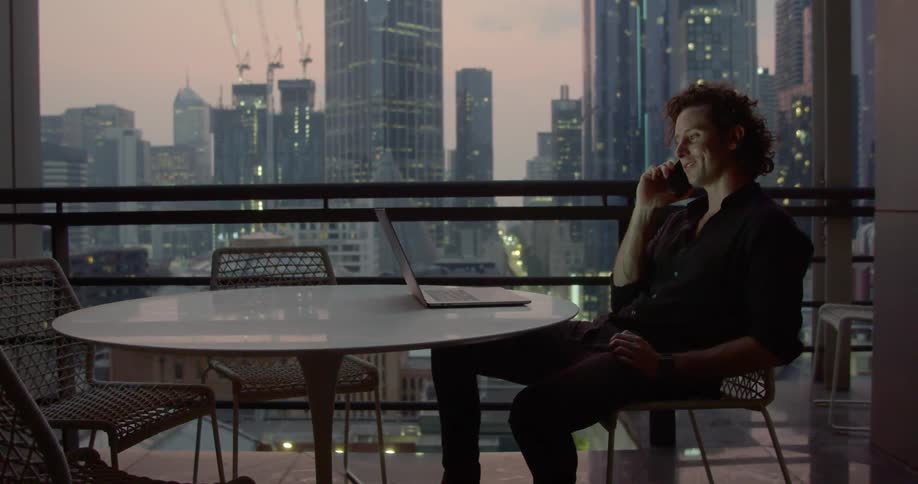 A man talking on his cell phone sits at a table next to a large window with a view a metropolitan area in the background