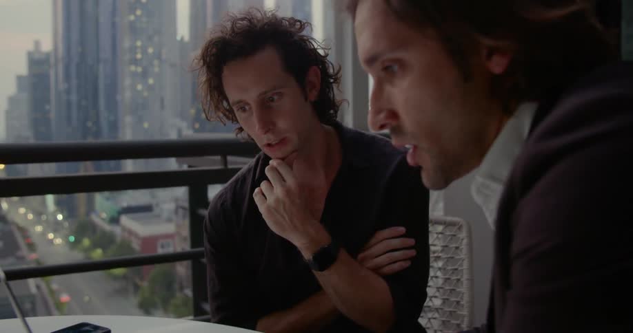 Two men sit and talk and look at a laptop