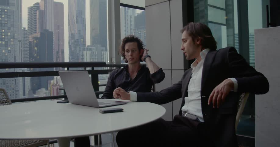 Two men sit at a table with a laptop beside large business office windows