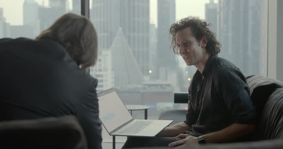 Two men sit together with a laptop beside large office windows in a city building