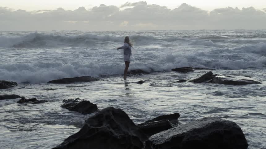 A woman walking into waves on a rocky beach