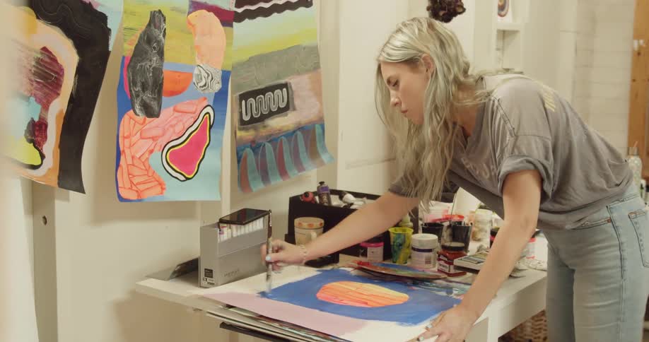 A woman stands at a table with artwork hanging around and uses a brush to paint blue on paper