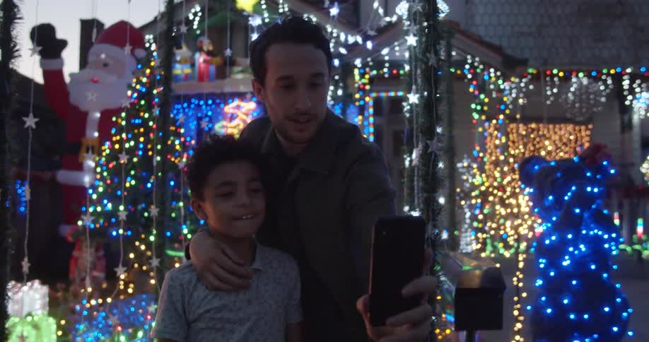 A dad takes a selfie of himself and son in front of a colorful christmas light display