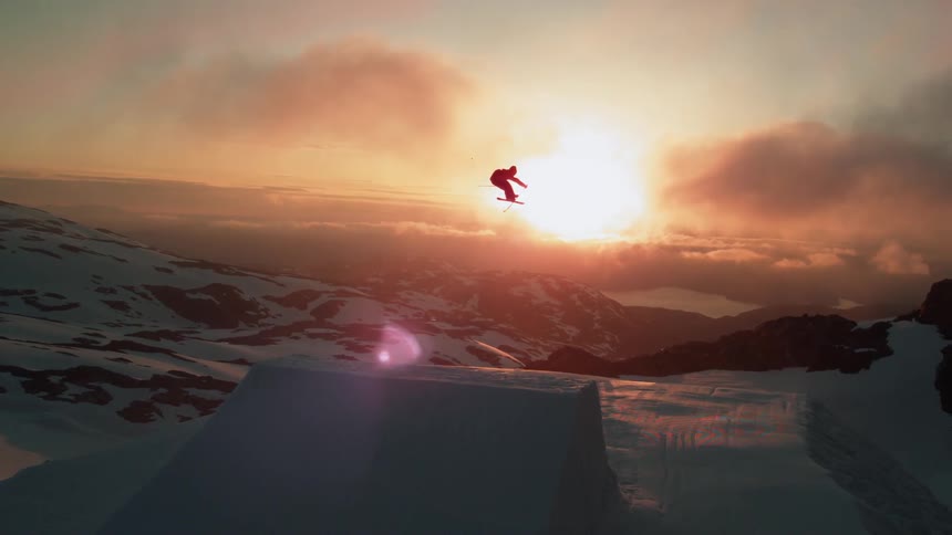 A skier spins in the air after going off a jump in snowy mountains at sunset