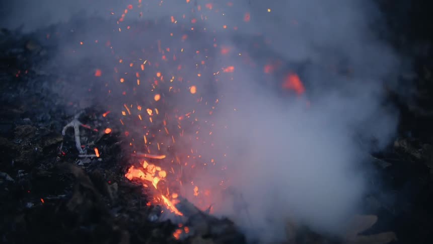 Close up of fire and smoking pieces of wood while parks fly in the night sky