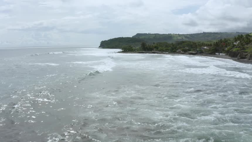 Broad ocean waves sweep towards a rocky coastline lined with many trees