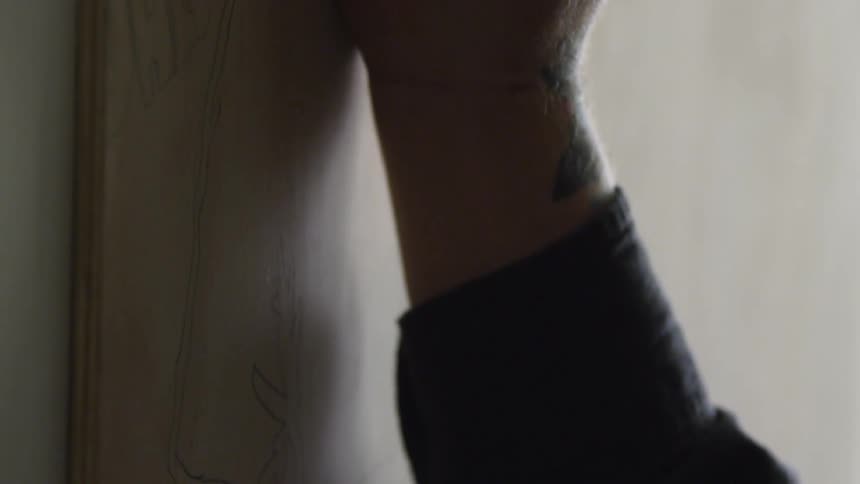 A man's arm rests upon a wooden board while he paints over line drawings
